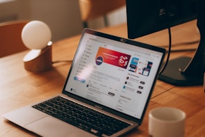 A laptop on a wooden desk displaying a social media page. The screen shows content in a web browser with various colors and text. A cup of coffee or tea sits beside the laptop, along with a modern lamp featuring a spherical bulb. In the background, there is a monitor partially visible.
