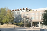 A modern, multi-story academic building with large glass windows and a distinctive roof design. Trees with green and autumn-colored leaves are in the foreground, and a paved walkway leads to the entrance. A few people are walking near the entrance, casting long shadows in the sunlight.