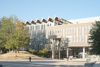 A modern, multi-story academic building with large glass windows and a distinctive roof design. Trees with green and autumn-colored leaves are in the foreground, and a paved walkway leads to the entrance. A few people are walking near the entrance, casting long shadows in the sunlight.