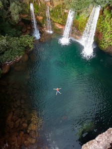 A serene natural pool features a person floating peacefully on their back in the center, surrounded by lush greenery and multiple cascading waterfalls. The surrounding rocks and trees add to the tranquil and natural setting.