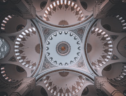 An intricately designed ceiling of a mosque with ornate geometric patterns and calligraphy encircling a large central dome. Soft light filters through small, arched windows, enhancing the symmetrical and detailed architectural features.