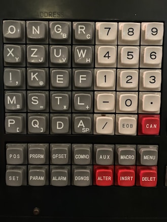 A vintage control panel featuring an array of buttons with letters, numbers, and commands. The buttons are arranged in rows and columns, with some buttons labeled with single characters and others with commands like POS, PRGRM, and AUX. Notably, there are several red buttons, including CAN, ALTER, and DELETE. The overall layout is reminiscent of an old computer or machinery interface.