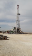 A tall drilling rig stands in an open, gravel-covered area with a partly cloudy sky overhead. The rig is surrounded by various machinery and equipment. The scene is expansive and industrial, with a sense of openness.