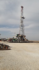 A tall drilling rig stands in an open, gravel-covered area with a partly cloudy sky overhead. The rig is surrounded by various machinery and equipment. The scene is expansive and industrial, with a sense of openness.