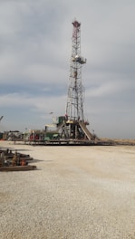 A tall drilling rig stands in an open, gravel-covered area with a partly cloudy sky overhead. The rig is surrounded by various machinery and equipment. The scene is expansive and industrial, with a sense of openness.