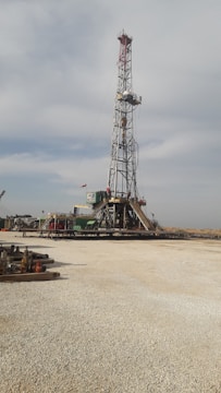 A tall drilling rig stands in an open, gravel-covered area with a partly cloudy sky overhead. The rig is surrounded by various machinery and equipment. The scene is expansive and industrial, with a sense of openness.