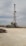 A tall drilling rig stands in an open, gravel-covered area with a partly cloudy sky overhead. The rig is surrounded by various machinery and equipment. The scene is expansive and industrial, with a sense of openness.