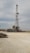 A tall drilling rig stands in an open, gravel-covered area with a partly cloudy sky overhead. The rig is surrounded by various machinery and equipment. The scene is expansive and industrial, with a sense of openness.