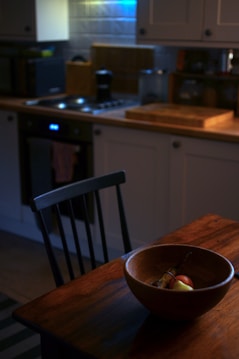 A cozy kitchen setting with warm lighting features a wooden table in the foreground and a bowl of fruit including apples and bananas. In the background, kitchen appliances like an oven, cutting boards, and a coffee maker are visible against tiled walls.