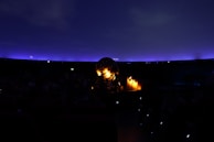 A dark interior scene featuring a planetarium with a glowing spherical projector at the center. The ceiling resembles a night sky with a gradient of deep blue to purple, dotted subtly with small points of light. Some seats and people are faintly visible in the shadowy environment.