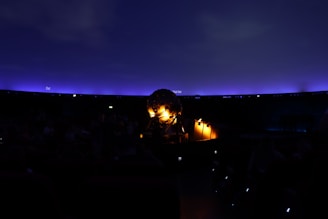 A dark interior scene featuring a planetarium with a glowing spherical projector at the center. The ceiling resembles a night sky with a gradient of deep blue to purple, dotted subtly with small points of light. Some seats and people are faintly visible in the shadowy environment.