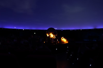 A dark interior scene featuring a planetarium with a glowing spherical projector at the center. The ceiling resembles a night sky with a gradient of deep blue to purple, dotted subtly with small points of light. Some seats and people are faintly visible in the shadowy environment.