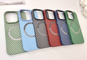 A collection of six carbon fiber-patterned phone cases, each with a circular magnetic ring design. The cases are arranged in a cascading order, showcasing a variety of colors including green, light blue, black, red, dark blue, and another shade of green.