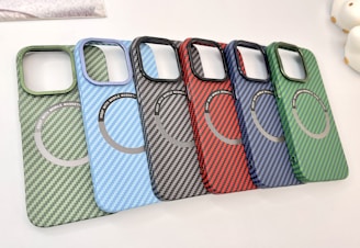A collection of six carbon fiber-patterned phone cases, each with a circular magnetic ring design. The cases are arranged in a cascading order, showcasing a variety of colors including green, light blue, black, red, dark blue, and another shade of green.