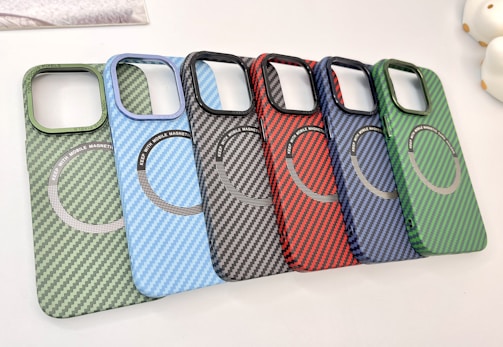A collection of six carbon fiber-patterned phone cases, each with a circular magnetic ring design. The cases are arranged in a cascading order, showcasing a variety of colors including green, light blue, black, red, dark blue, and another shade of green.