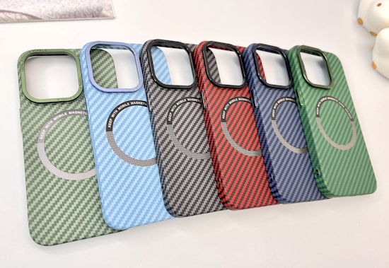 A collection of six carbon fiber-patterned phone cases, each with a circular magnetic ring design. The cases are arranged in a cascading order, showcasing a variety of colors including green, light blue, black, red, dark blue, and another shade of green.