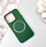 A green and black striped phone case with a cutout for the camera and a magnetic card holder indication. Two rabbit-shaped white objects are placed to the right, and a subtle floral design is in the background.