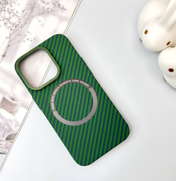 A green and black striped phone case with a cutout for the camera and a magnetic card holder indication. Two rabbit-shaped white objects are placed to the right, and a subtle floral design is in the background.