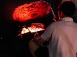 A person cooks food using an open flame, with a grill positioned over the fire. The ambiance is dark, with orange and red hues illuminating the area, while a warm glow emanates from the flames.