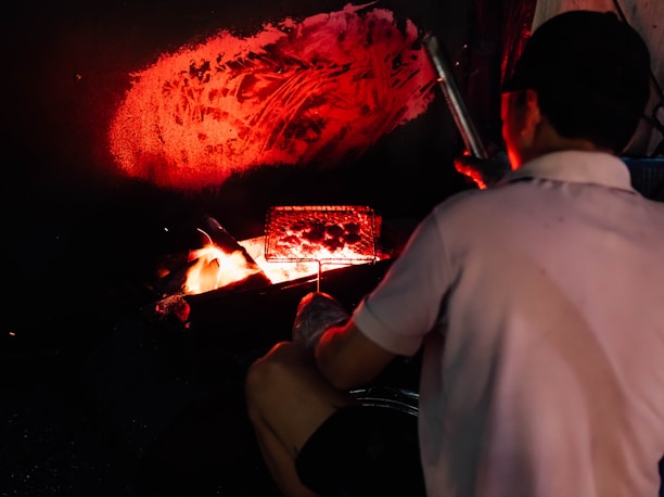 A person cooks food using an open flame, with a grill positioned over the fire. The ambiance is dark, with orange and red hues illuminating the area, while a warm glow emanates from the flames.