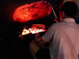 A person cooks food using an open flame, with a grill positioned over the fire. The ambiance is dark, with orange and red hues illuminating the area, while a warm glow emanates from the flames.