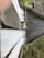 Aerial view of a dam with water cascading down the spillway. The structure is made of concrete with a series of interlocking blocks. Surrounding the dam is lush green grass and pathways. The water creates white foamy textures as it falls.