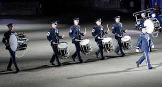 A group of uniformed individuals, likely a military marching band, playing large drums in an organized formation. The drummers are wearing dark blue uniforms with matching caps, and they appear to be participating in a ceremonial or formal event. The setting is likely outdoors on a flat surface, possibly during the evening or night.