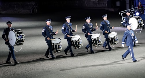A group of uniformed individuals, likely a military marching band, playing large drums in an organized formation. The drummers are wearing dark blue uniforms with matching caps, and they appear to be participating in a ceremonial or formal event. The setting is likely outdoors on a flat surface, possibly during the evening or night.