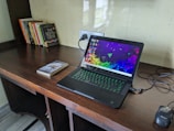 A wooden desk featuring a laptop with a colorful screen saver and a glowing keyboard. Nearby lies a black optical mouse and a few books, including a prominent one with the title 'The Selfish Gene'. The wall behind is light-colored with a reflective surface and there's an electrical socket in view.
