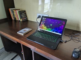 A wooden desk featuring a laptop with a colorful screen saver and a glowing keyboard. Nearby lies a black optical mouse and a few books, including a prominent one with the title 'The Selfish Gene'. The wall behind is light-colored with a reflective surface and there's an electrical socket in view.