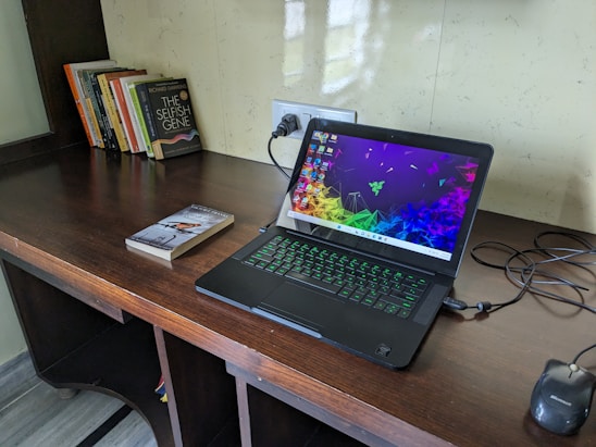 A wooden desk featuring a laptop with a colorful screen saver and a glowing keyboard. Nearby lies a black optical mouse and a few books, including a prominent one with the title 'The Selfish Gene'. The wall behind is light-colored with a reflective surface and there's an electrical socket in view.