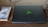 A closed black laptop with a green logo is placed on a dark wooden table. Next to it lies a paperback book titled 'Man's Search for Meaning' by Viktor E. Frankl. The book cover features a small bird and barbed wire, hinting at a somber theme.