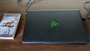 A closed black laptop with a green logo is placed on a dark wooden table. Next to it lies a paperback book titled 'Man's Search for Meaning' by Viktor E. Frankl. The book cover features a small bird and barbed wire, hinting at a somber theme.