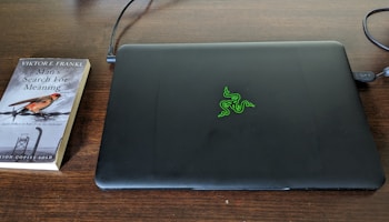 A closed black laptop with a green logo is placed on a dark wooden table. Next to it lies a paperback book titled 'Man's Search for Meaning' by Viktor E. Frankl. The book cover features a small bird and barbed wire, hinting at a somber theme.