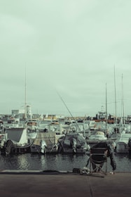 A fisherman sits on a folding chair with a fishing rod extended towards the water. In the background, numerous boats are moored at a harbor, covered with tarpaulins. The overall atmosphere is tranquil, with muted tones and a slightly overcast sky.