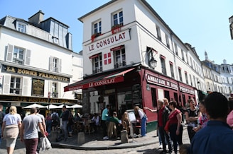 A bustling street scene in a historic district with cafes and restaurants. The central building has 'Le Consulat' on its facade, decorated with red accents and flower boxes on the windows. People are gathered around, enjoying the lively atmosphere beneath a clear blue sky. The architecture features classic European style with narrow streets and vibrant signage.