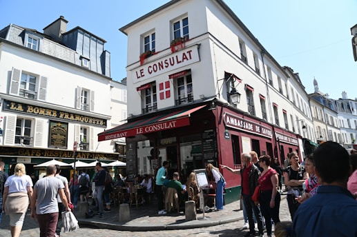 A bustling street scene in a historic district with cafes and restaurants. The central building has 'Le Consulat' on its facade, decorated with red accents and flower boxes on the windows. People are gathered around, enjoying the lively atmosphere beneath a clear blue sky. The architecture features classic European style with narrow streets and vibrant signage.