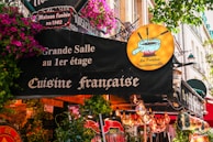 A lively French restaurant storefront adorned with colorful flowers and quaint signage. The sign advertises traditional French cuisine, specifically fondue, with a whimsical illustration of a fondue pot. The exterior is vibrant with hanging lights and various decorative elements.