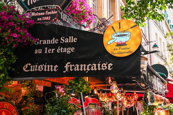 A lively French restaurant storefront adorned with colorful flowers and quaint signage. The sign advertises traditional French cuisine, specifically fondue, with a whimsical illustration of a fondue pot. The exterior is vibrant with hanging lights and various decorative elements.
