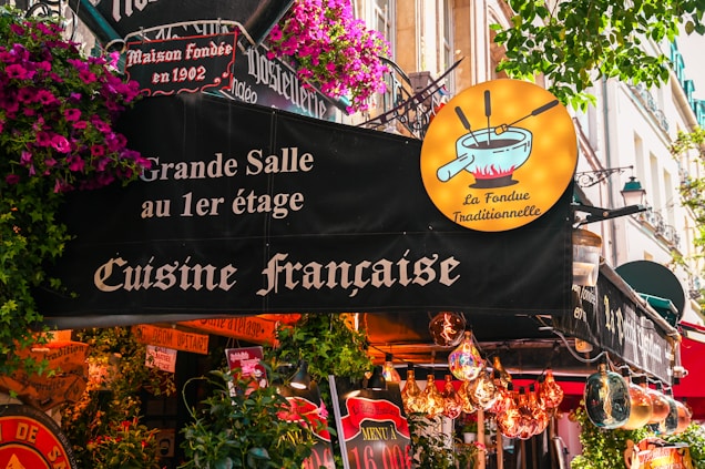 A lively French restaurant storefront adorned with colorful flowers and quaint signage. The sign advertises traditional French cuisine, specifically fondue, with a whimsical illustration of a fondue pot. The exterior is vibrant with hanging lights and various decorative elements.