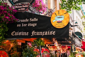 A lively French restaurant storefront adorned with colorful flowers and quaint signage. The sign advertises traditional French cuisine, specifically fondue, with a whimsical illustration of a fondue pot. The exterior is vibrant with hanging lights and various decorative elements.