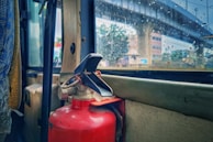 A red fire extinguisher is mounted inside a vehicle next to a rain-speckled window, through which an urban scene with a bridge and buildings is visible. The interior appears to have worn surfaces and the window reveals raindrops, suggesting rainy weather outside.
