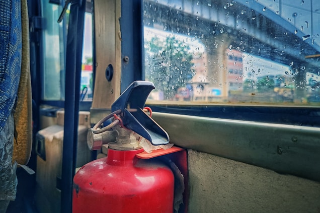 A red fire extinguisher is mounted inside a vehicle next to a rain-speckled window, through which an urban scene with a bridge and buildings is visible. The interior appears to have worn surfaces and the window reveals raindrops, suggesting rainy weather outside.