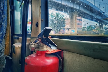 A red fire extinguisher is mounted inside a vehicle next to a rain-speckled window, through which an urban scene with a bridge and buildings is visible. The interior appears to have worn surfaces and the window reveals raindrops, suggesting rainy weather outside.