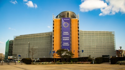 The image features a large, modern building with a distinct angular design, partially covered by a banner displaying the words 'Europese Commissie', 'Commission européenne', and 'European Commission' along with the EU emblem. The building has a prominent glass facade and is situated under a clear blue sky with scattered clouds. Flags are visible near the base, and there are some trees and shrubs in front of the building.