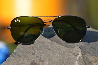 A pair of aviator-style sunglasses with dark green lenses rests on a rough, textured rock surface. The metal frame glimmers in the light, and a soft, blurred background featuring warm orange and yellow tones suggests a sunset or sunrise.