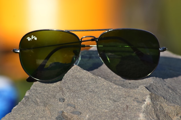 A pair of aviator-style sunglasses with dark green lenses rests on a rough, textured rock surface. The metal frame glimmers in the light, and a soft, blurred background featuring warm orange and yellow tones suggests a sunset or sunrise.