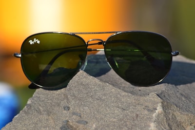 A pair of aviator-style sunglasses with dark green lenses rests on a rough, textured rock surface. The metal frame glimmers in the light, and a soft, blurred background featuring warm orange and yellow tones suggests a sunset or sunrise.