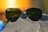 A pair of aviator-style sunglasses with dark green lenses rests on a rough, textured rock surface. The metal frame glimmers in the light, and a soft, blurred background featuring warm orange and yellow tones suggests a sunset or sunrise.
