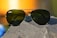 A pair of aviator-style sunglasses with dark green lenses rests on a rough, textured rock surface. The metal frame glimmers in the light, and a soft, blurred background featuring warm orange and yellow tones suggests a sunset or sunrise.